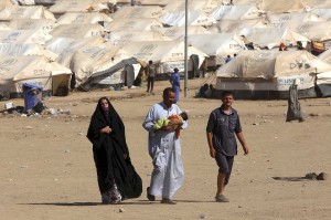 A woman and two men, one of whom is holding an infant, walk on a dry dusty road. Hundreds of refugee tents are behind them.