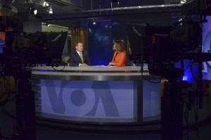 Two anchors sit at a VOA news desk.