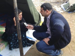 Male MBN reporter kneels in front of a tent, interviewing a displaced woman.