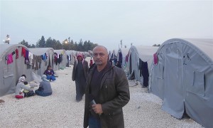 A reporter stands in front of a refugee camp -several tents with people milling about are behind him.