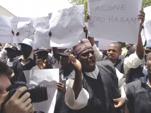 Several men, some wearing traditional hats, some wearing baseball0style caps, protest against Boko Haram. The men are carrying signs that read, "#bringbackourgirls safe and alive" and "Our hearts are with the parents."