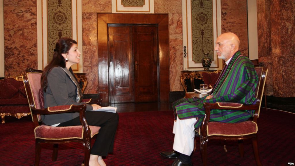 Female reporter sits across from President Karzai who is dressed in green and purple traditional dress. They sit in an ornately decorated room.